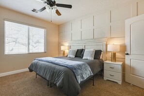 Une chambre à coucher avec un lit, des tables de chevet, un ventilateur de plafond et une fenêtre avec des stores.