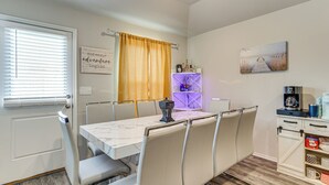 Un coin repas avec une table et des chaises, une fenêtre avec des stores, un rideau, une étagère fixée au mur avec quelques objets, et un tableau accroché au mur.