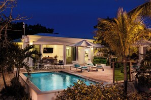 Un'area piscina moderna con poltrone prendisole, una piscina e una casa ben illuminata sullo sfondo.