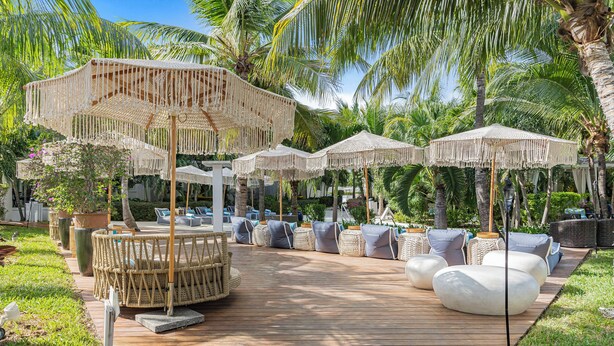 Une terrasse en bois avec des espaces de détente en plein air, comprenant des tables et des chaises rondes, le tout entouré de palmiers et d’ombrelles.
