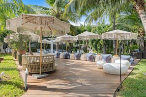 A wooden deck with outdoor seating, including round tables and chairs, surrounded by palm trees and umbrellas. - Deluxe King Room Studio (Providenciales)