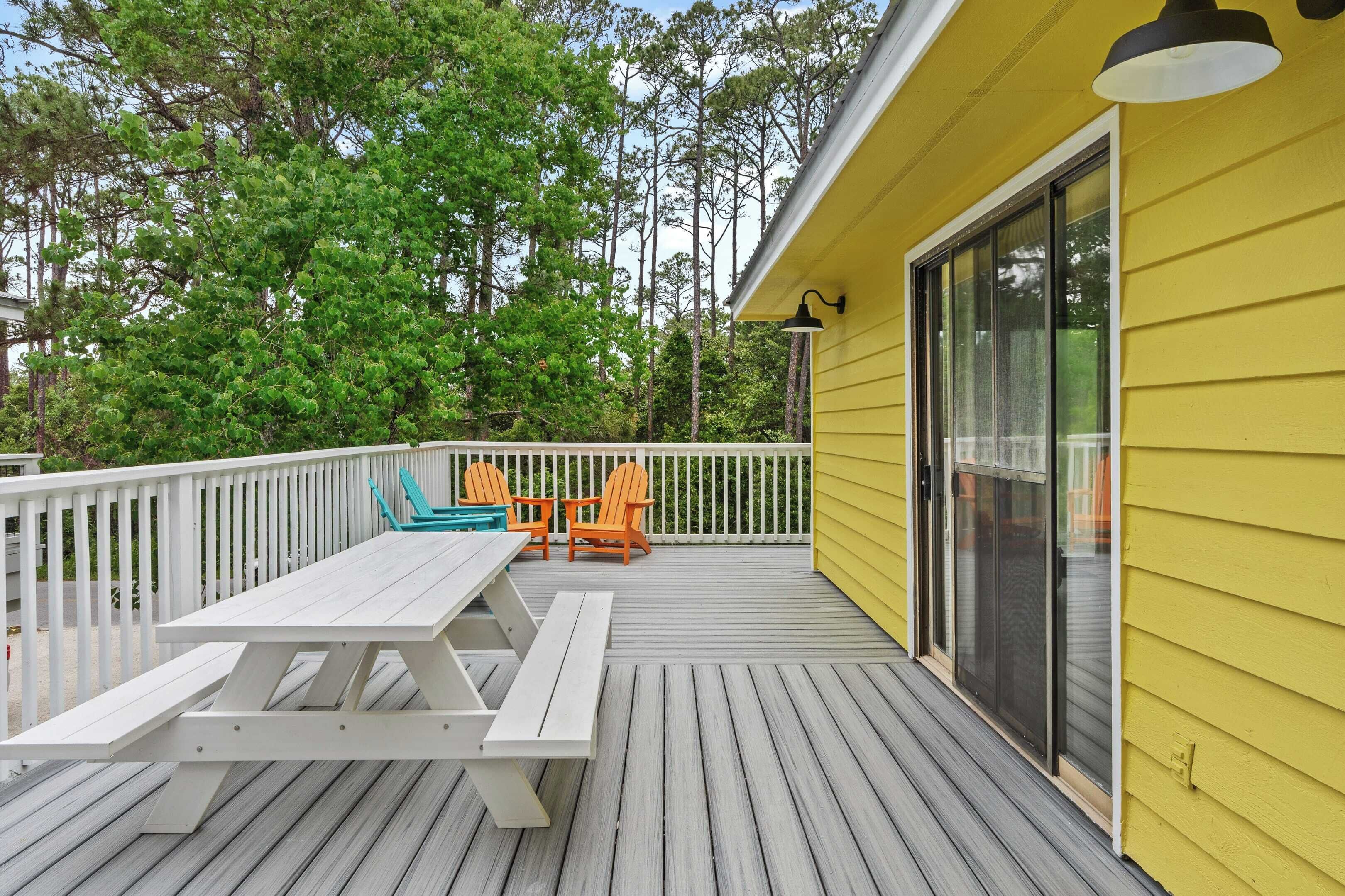 A wooden deck with a picnic table and chairs, a yellow building with a sliding glass door, and a white fence.