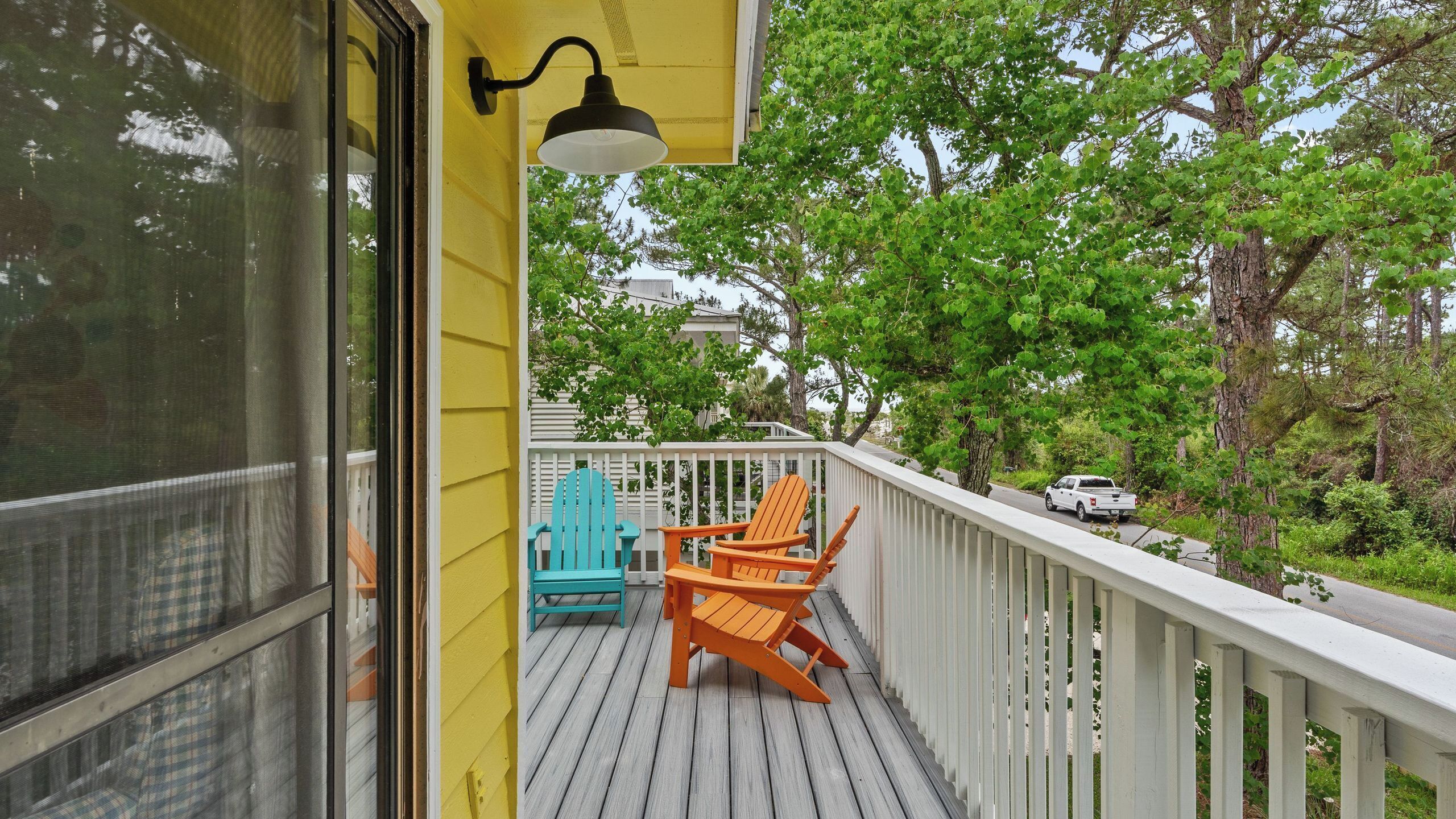 Une terrasse en bois avec deux fauteuils Adirondack, un camionnette blanche et un bâtiment jaune doté d’un porche grillagé.