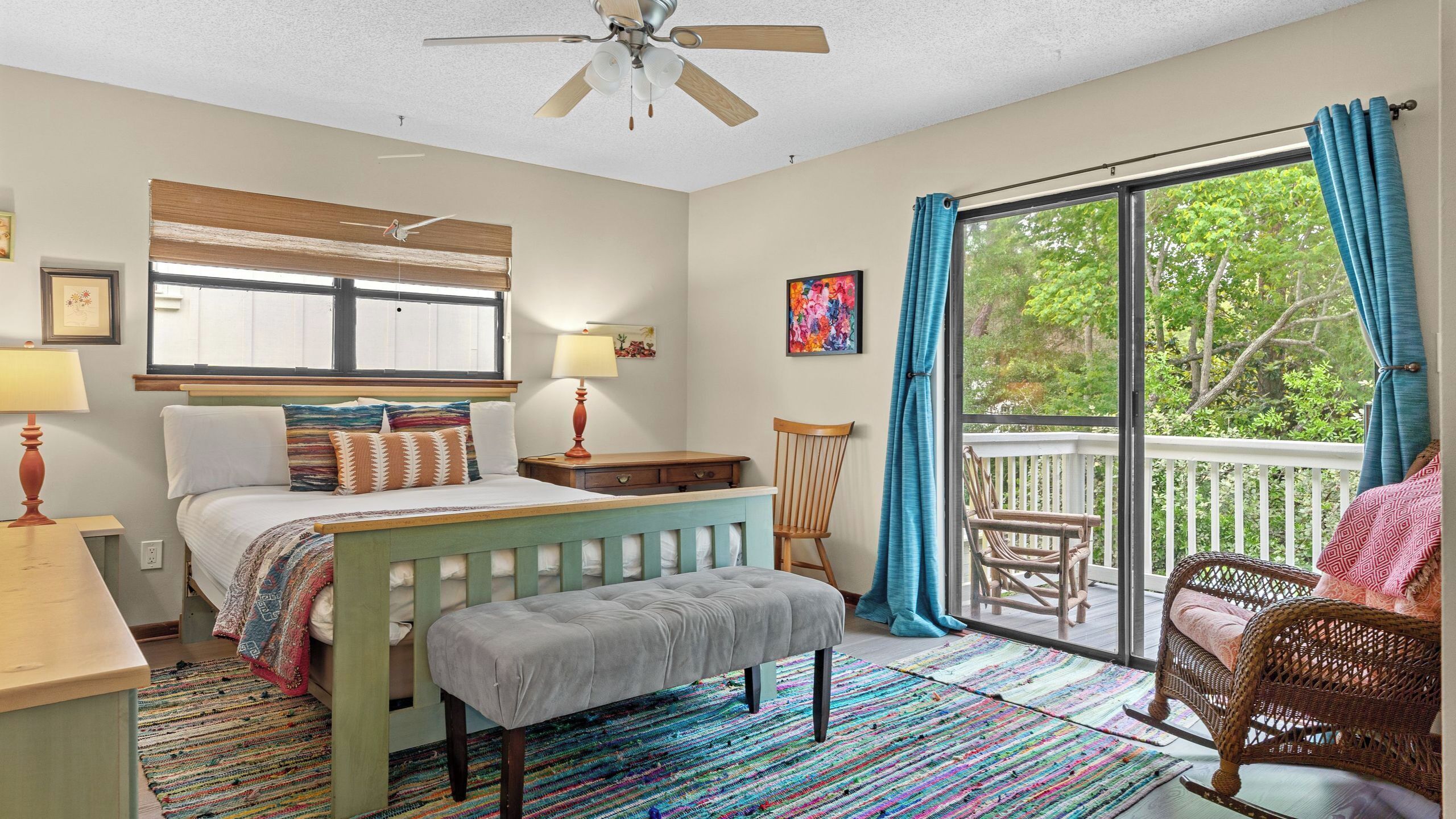 A bedroom with a bed, bedside table, lamp, chair, bench, and a view of the outdoors through a sliding glass door.