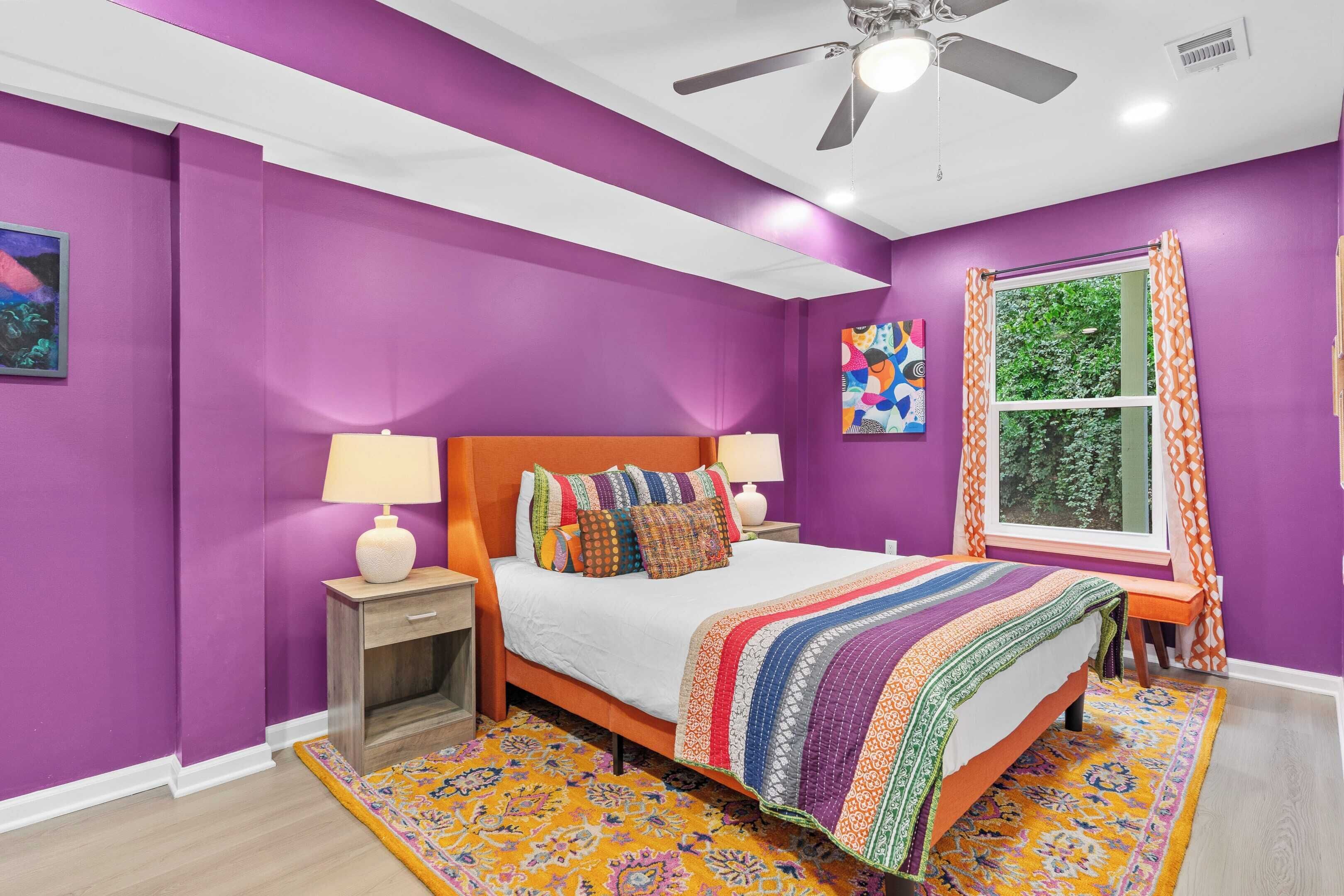 Un dormitorio con paredes moradas, una cama de madera, un tapete colorido y una ventana con cortinas.