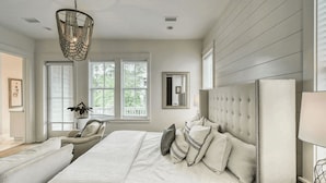 A bedroom with a large bed, a chair, a chandelier, and a window with blinds.