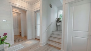 A hallway with white walls and wooden floors, leading to a staircase with a potted plant on the landing.