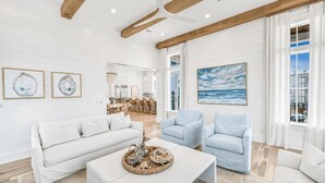 Un salon spacieux avec un canapé blanc, des fauteuils bleu clair et une table basse agrémentée d’objets décoratifs. Un grand tableau représentant l’océan orne le mur, et un coin repas se trouve à l’arrière-plan.