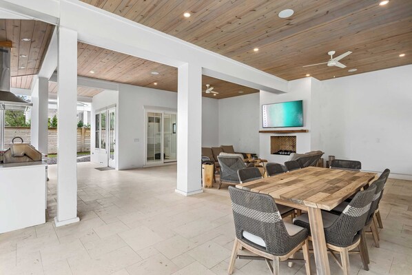 A spacious indoor area with a wooden ceiling, a large dining table with wicker chairs, a flat-screen TV mounted on the wall, and a view of the outdoors through glass doors.