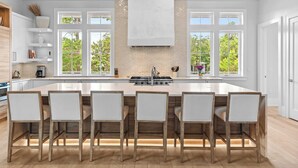 Een moderne keuken met een groot eiland, witte kasten en uitzicht op bomen door grote ramen.
