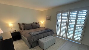 A bedroom with a large bed, two bedside tables, a nightstand, a bench, and a sliding glass door with blinds.