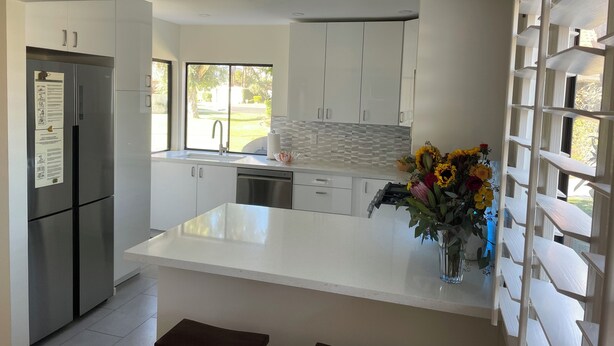 A modern kitchen with white cabinetry, a stainless steel refrigerator, a large island with a vase of flowers, and a view of the outdoors through a window.