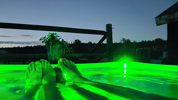 A person relaxing in a green-lit hot tub with a view of trees and a building in the background.