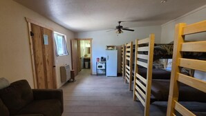 A room with bunk beds, a sofa, a small stool, a ceiling fan, and a refrigerator in the background.