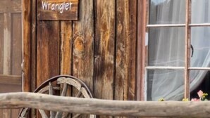 En rustik træbygning med et skilt der siger "Wrangler", et stort træhjul og et vindue med gardiner.