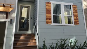 Une maison à deux étages avec une entrée à l’avant, un escalier en bois et un petit jardin agrémenté d’arbustes.