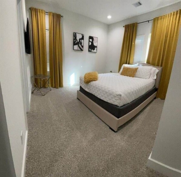 Un dormitorio con cama, cortinas, una mesita y dos cuadros.
