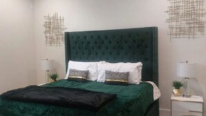 Une chambre à coucher avec un lit recouvert de velours vert, des oreillers blancs et une tête de lit vert foncé. On y trouve deux tables de chevet, chacune surmontée d’une lampe et d’une petite plante.