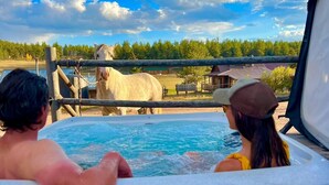 Un jacuzzi con dos personas adentro, con vistas a un área cercada donde hay un caballo y un edificio al fondo.