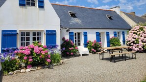 A white building with blue shutters and a sloped roof, surrounded by gravel and flowering plants. - Tipoz'Immo • Maison Bleue Walking distance to the beach • Surf spot • garden (Pouldreuzic)