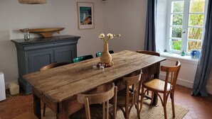A dining area with a wooden table, chairs, a vase with flowers, a wooden shelf, and a window with curtains. - Tipoz'Immo • Maison Bleue Walking distance to the beach • Surf spot • garden (Pouldreuzic)