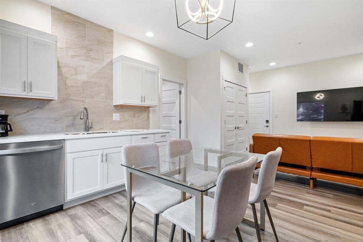 A modern kitchen with white cabinets, a stainless steel dishwasher, and a glass dining table with chairs.