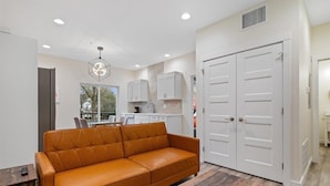A modern living room with a leather sofa, a dining table, and a kitchen area with white cabinets and a double door.
