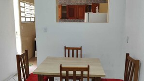 Un coin repas avec une table et des chaises en bois, une fenêtre ornée de rideaux à motifs et une cuisine visible à travers une porte ouverte.