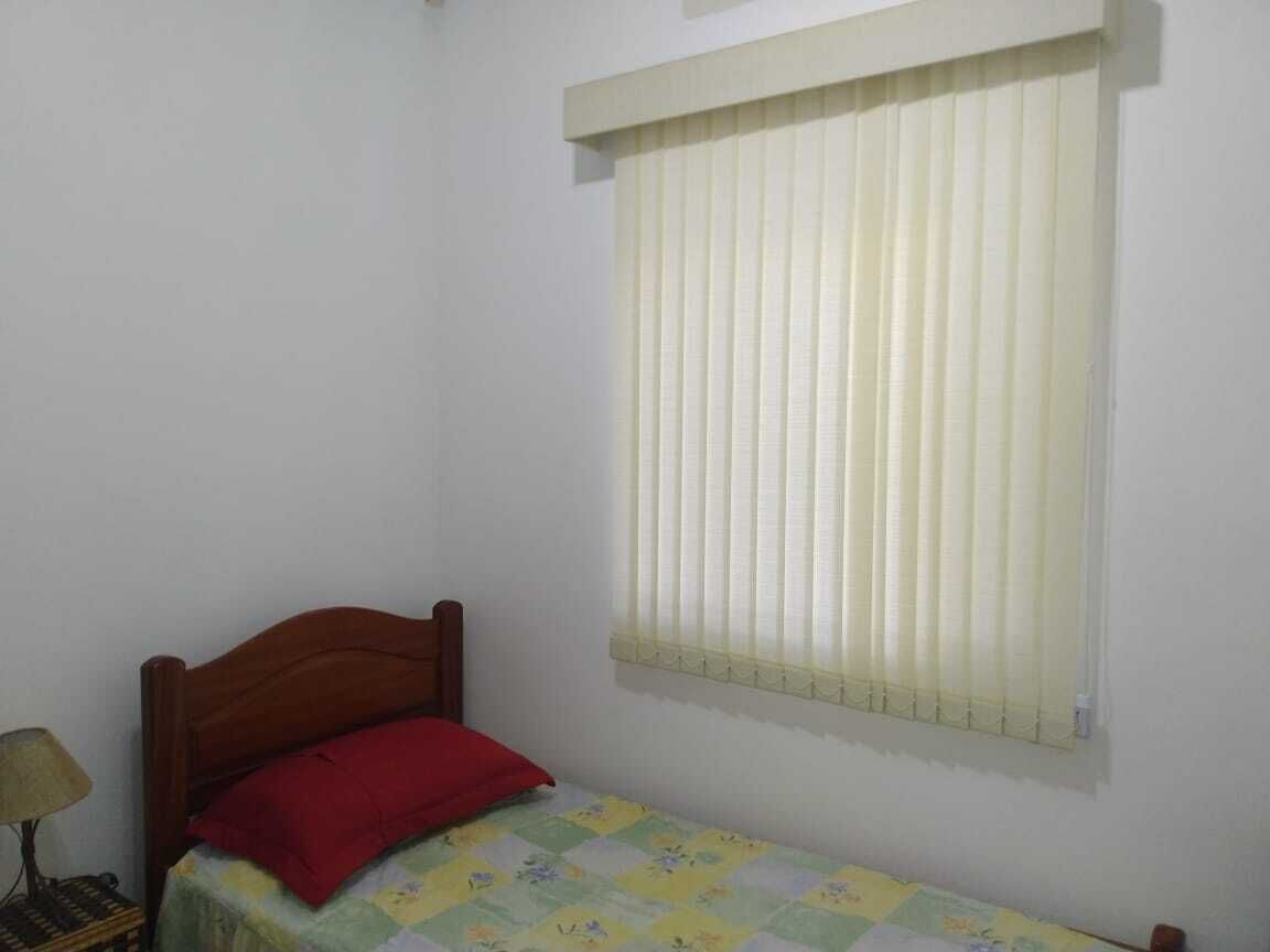Een kleine kamer met een enkel bed, een houten hoofdbord, een rood kussen en een raam met verticale jaloezieën.