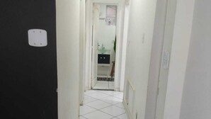 Un couloir étroit aux murs et au sol recouverts de carreaux blancs, qui mène à une salle de bain équipée d’un lavabo et d’un miroir.
