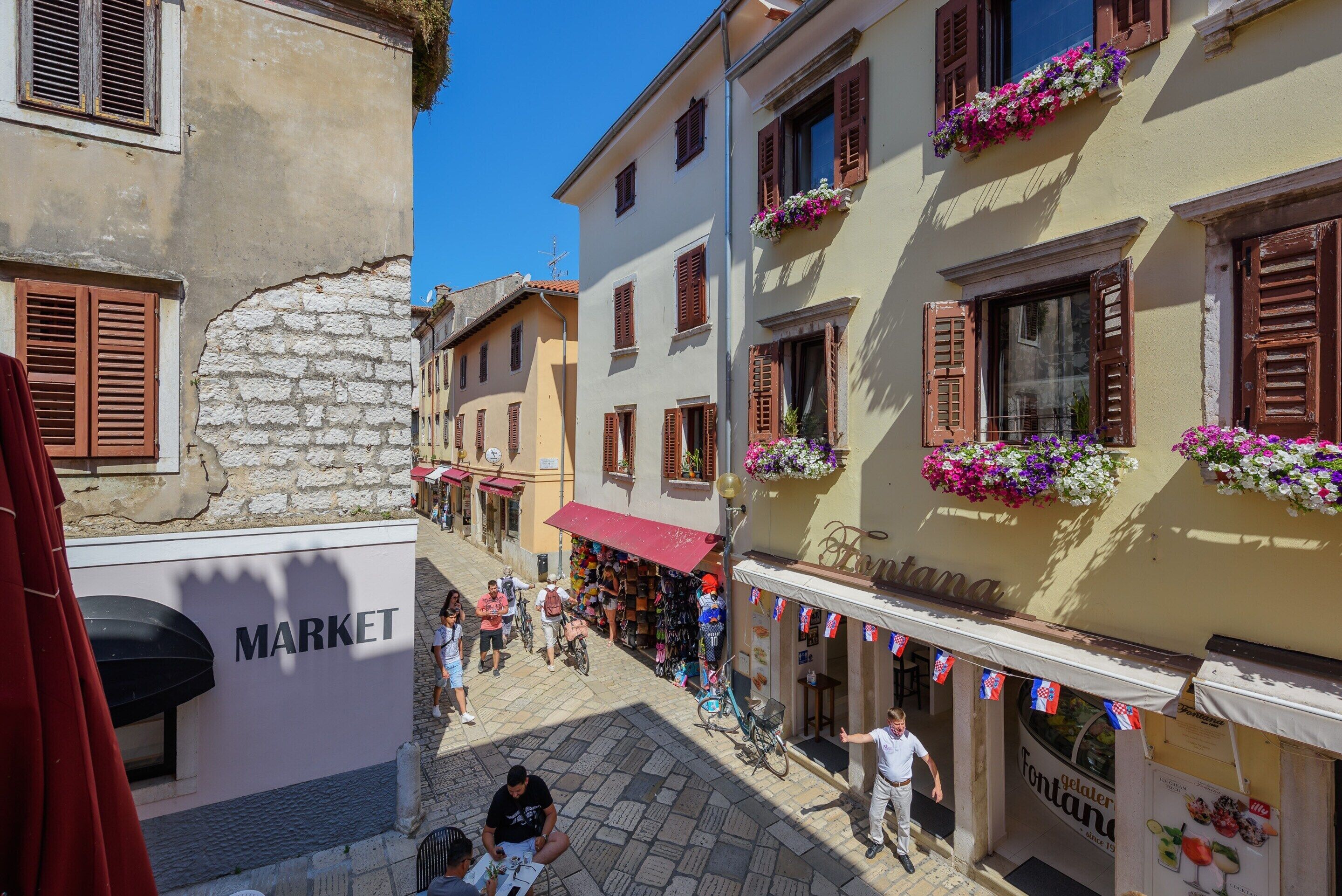 Une rue étroite bordée de bâtiments des deux côtés, une enseigne indiquant un marché et une devanture de magasin avec une pancarte sur laquelle on peut lire « Fontana ».