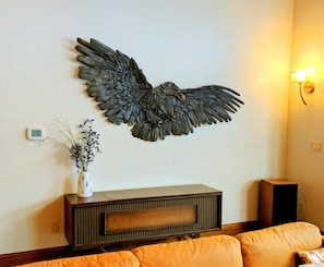 A room with a wall-mounted decorative bird sculpture, a small white vase with dried flowers, a wooden console table, and a brown leather sofa.