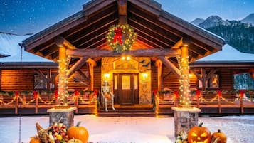 Une cabane en rondins, avec une façade en bois, des fondations en pierre et une véranda décorée de guirlandes lumineuses et d’une couronne.