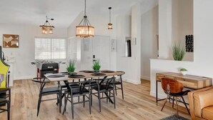 A spacious living area with a wooden dining table, black chairs, a sofa, and a coffee table. There is a kitchen area with a foosball table, and a large window with blinds. - 4BR Family Getaway w/Pool (North Las Vegas)