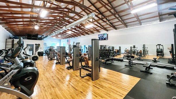 Een sportschool met diverse fitnessapparaten, houten vloeren en een hoge ruimte met zichtbare balken.