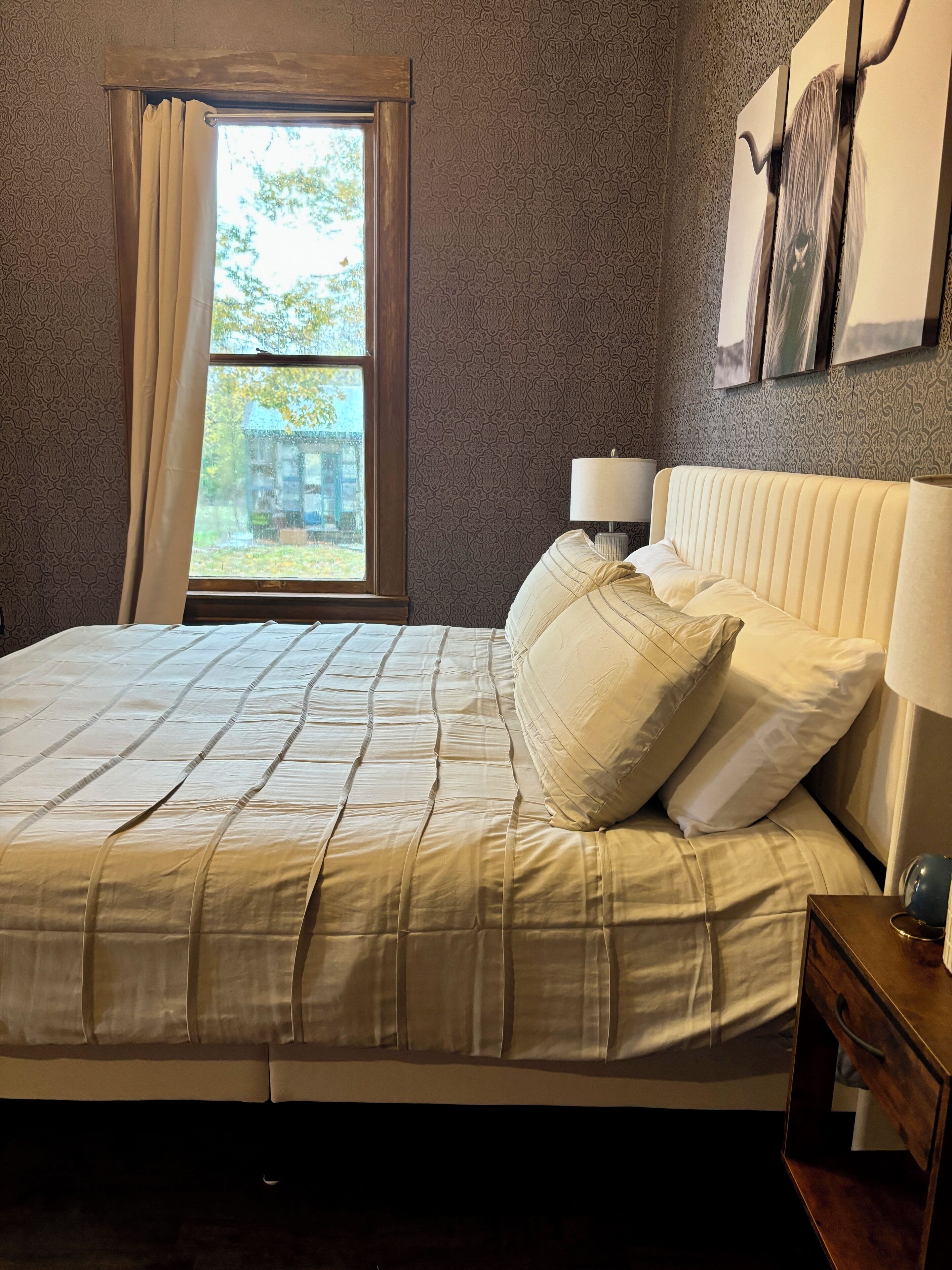 Une chambre à coucher avec un grand lit, une fenêtre avec des rideaux, une lampe de chevet et un mur orné de tableaux.
