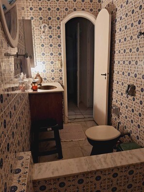 Un baño con baldosas estampadas, un tocador de madera, un lavabo, un inodoro y una puerta que da a otra habitación.
