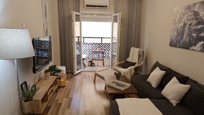 Ett modernt vardagsrum med en soffa, en fåtölj och ett soffbord. På vänster sida finns en tv, en krukväxt och ett fönster med gardiner.