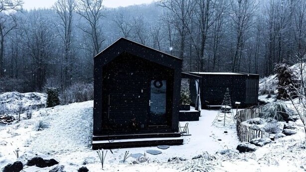 A cabin with a large window and a door, surrounded by a snowy landscape and trees.