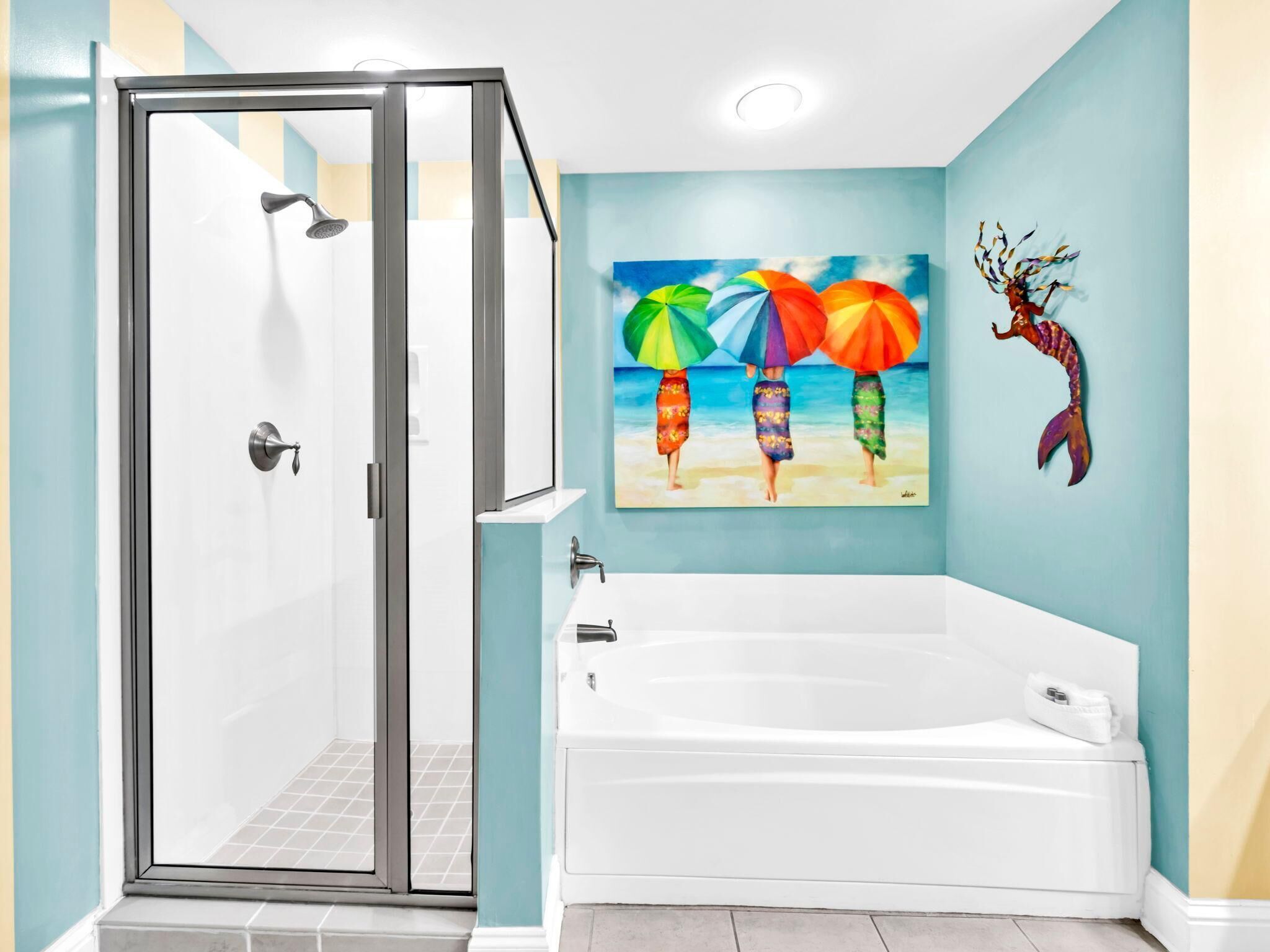 A bathroom with a glass-enclosed shower, a white bathtub, and a painting of beach umbrellas.