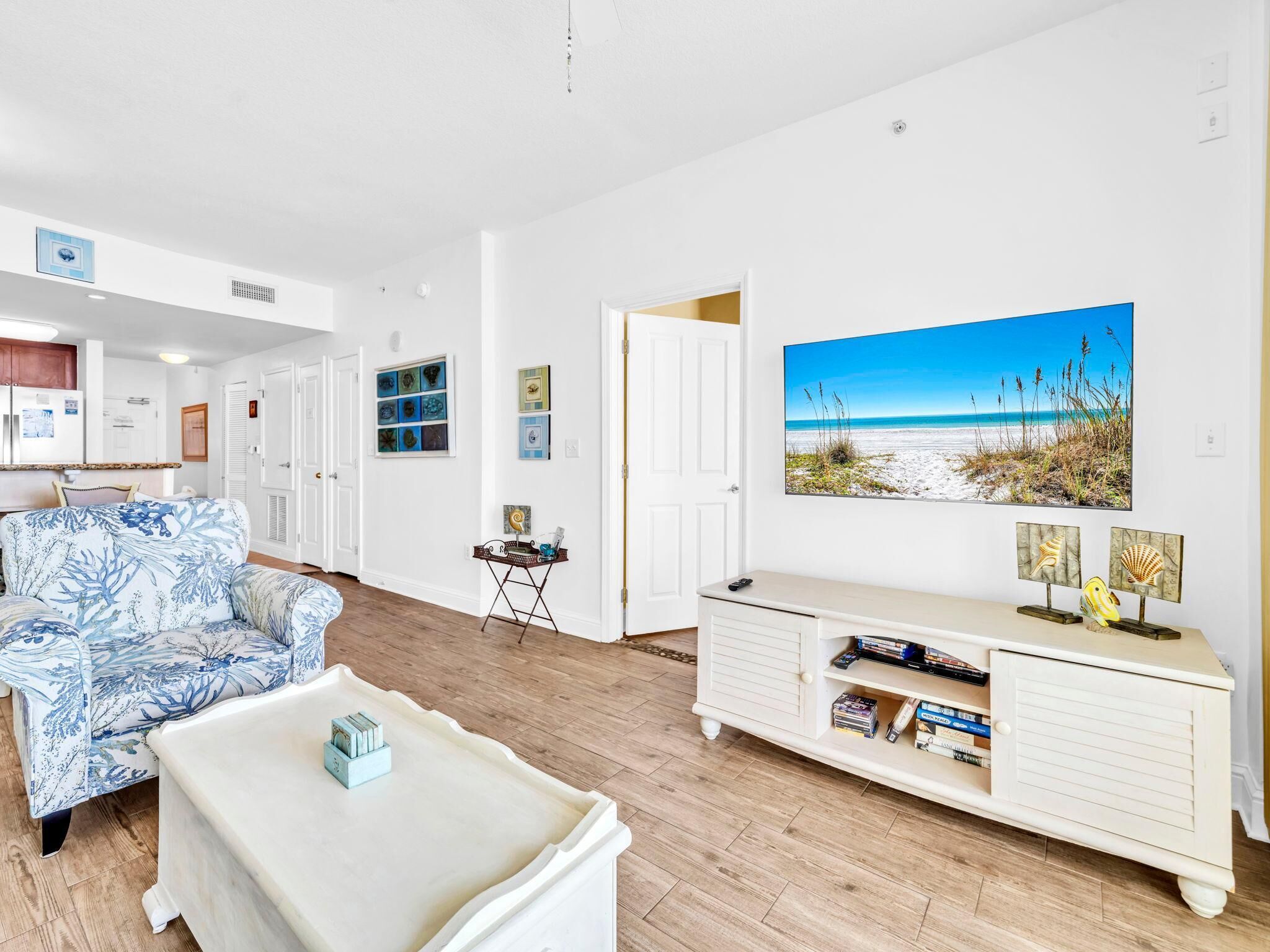Un salon spacieux avec un canapé blanc, une table basse et un meuble blanc rempli de livres. Un grand tableau représentant une plage orne le mur.