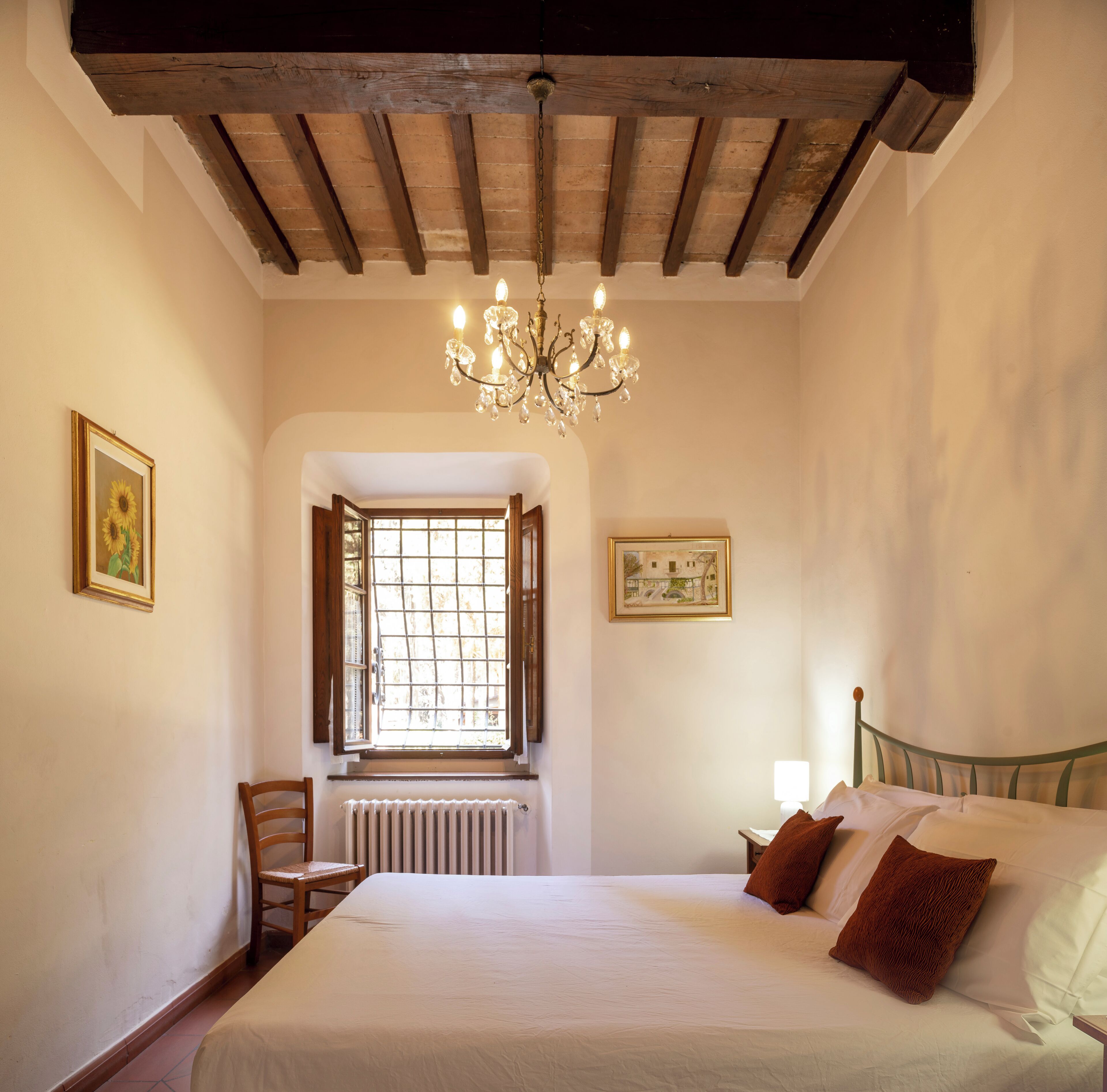 A bedroom with a large bed, a wooden chair, a radiator, a window with open shutters, and a chandelier.