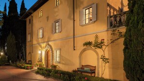 A two-story building with a warm facade, multiple windows with shutters, and a well-maintained garden with a bench. - Villa La Cappella  (Montespertoli)