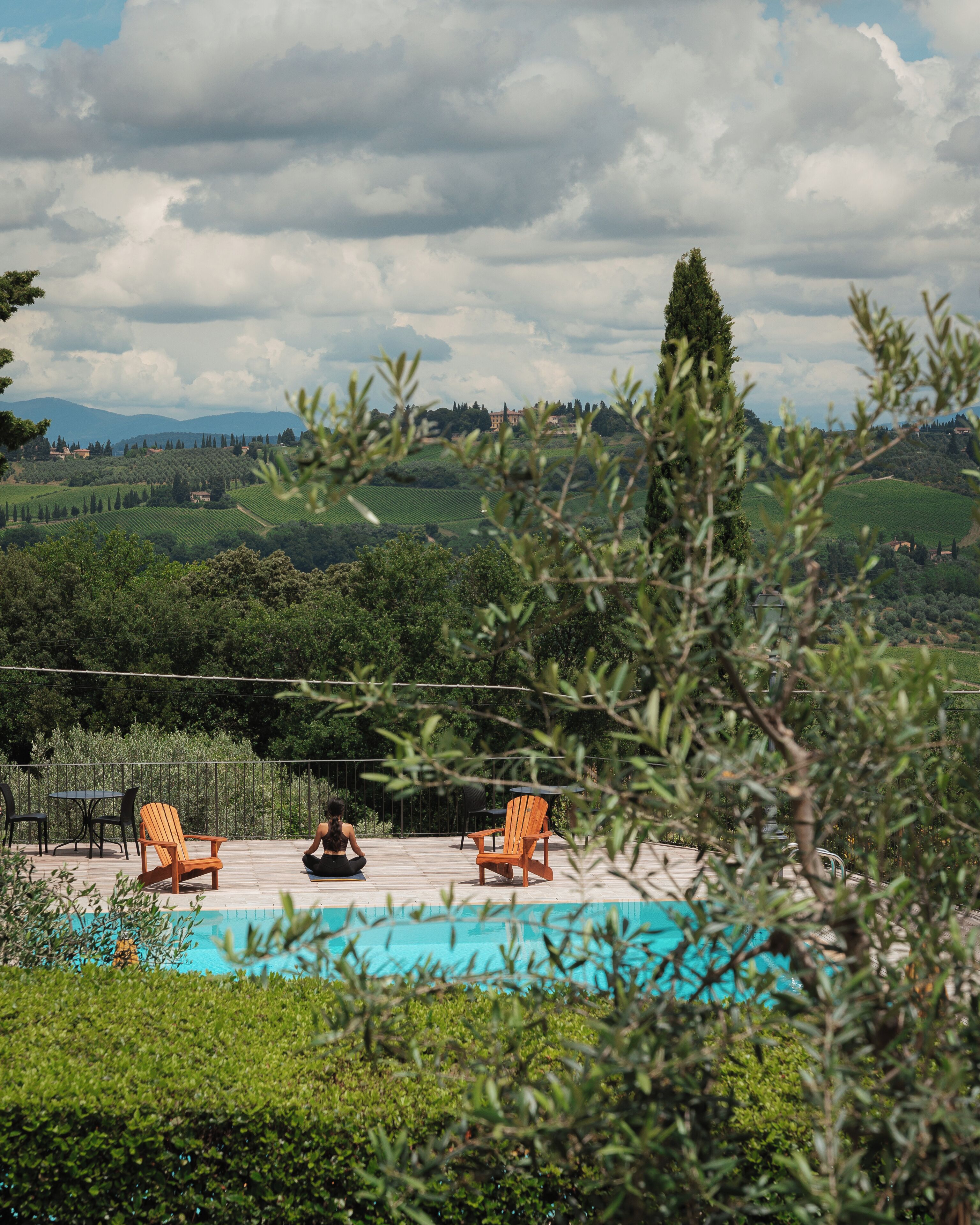 A pool area with two wooden chairs and a table, surrounded by lush greenery and a view of rolling hills.