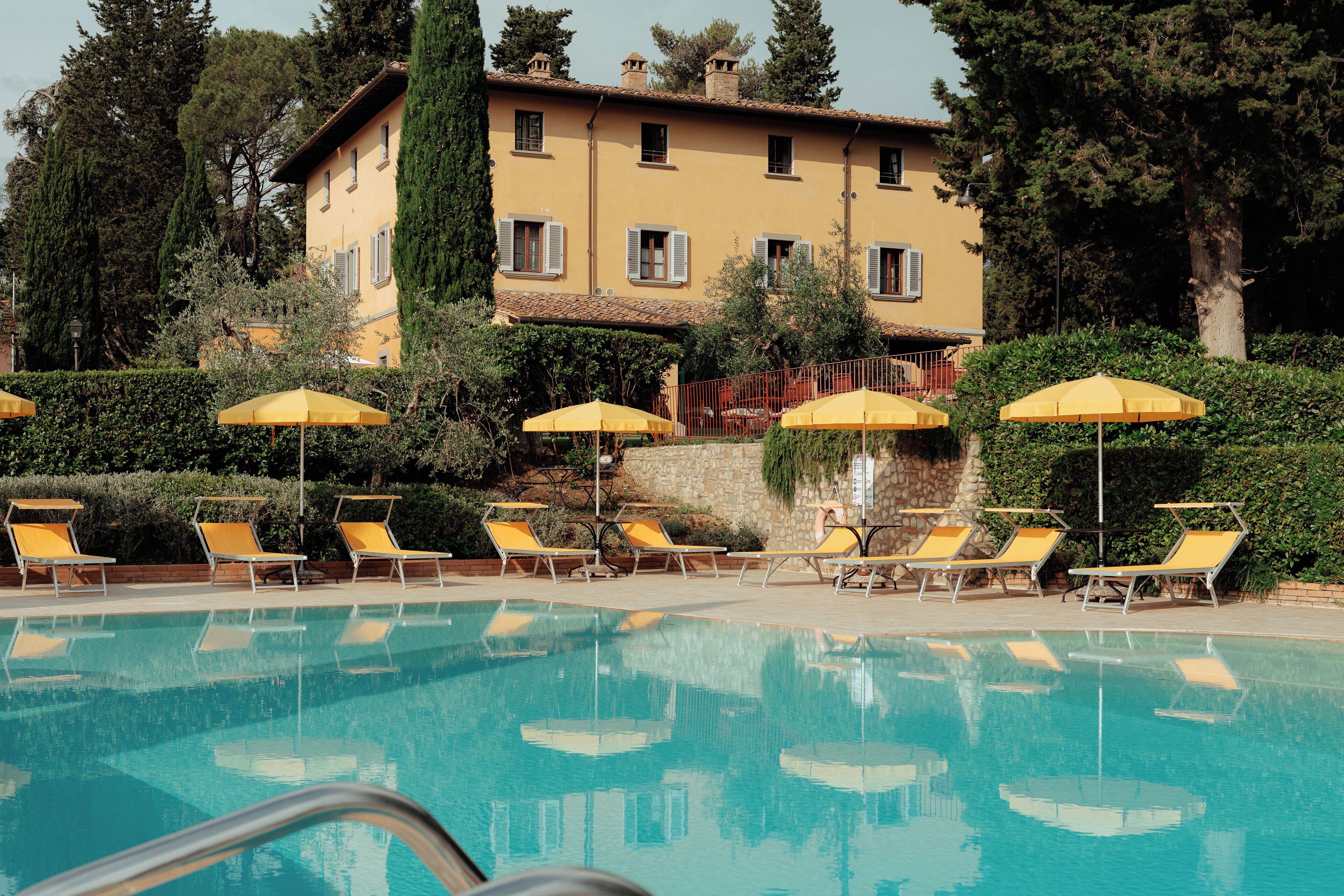 A pool area with sun loungers and umbrellas, a building with multiple windows and shutters, and surrounding greenery.