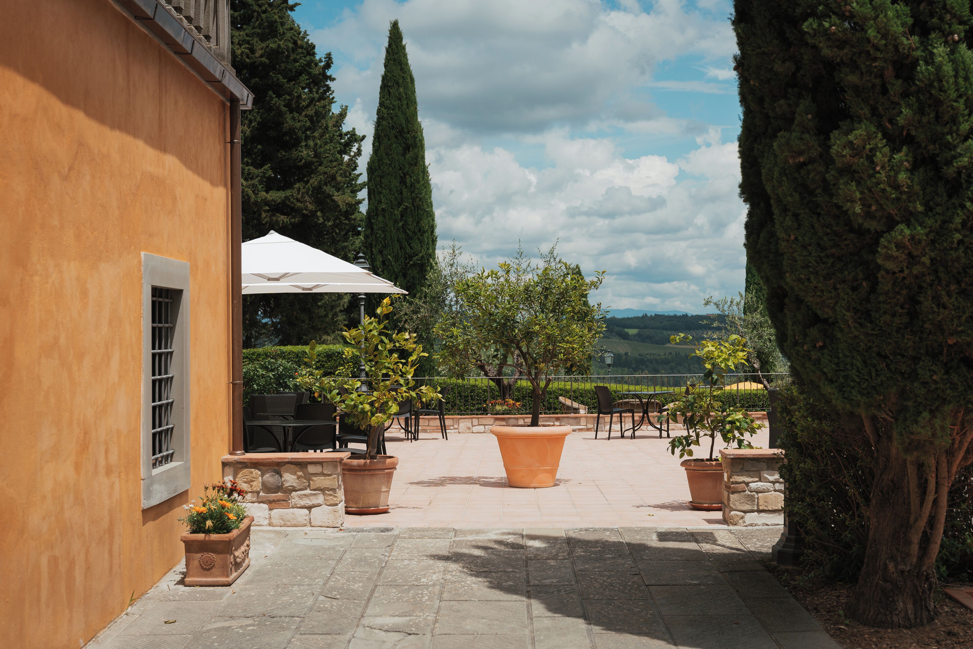 An outdoor patio with stone flooring, potted plants, a white umbrella, and a view of the countryside.