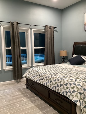 Quarto com cama, janela com cortinas, mesa de cabeceira com abajur e vista para uma varanda.