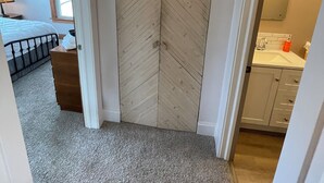 Un pasillo con puerta de madera, suelo alfombrado, cama con cabecera, cómoda y baño con lavabo y espejo.