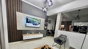 Un salon moderne avec un téléviseur, une table basse en bois, un canapé gris et un coin repas à l’arrière-plan.