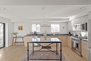 A modern kitchen with wooden cabinets, a central island, stainless steel appliances, and large windows.